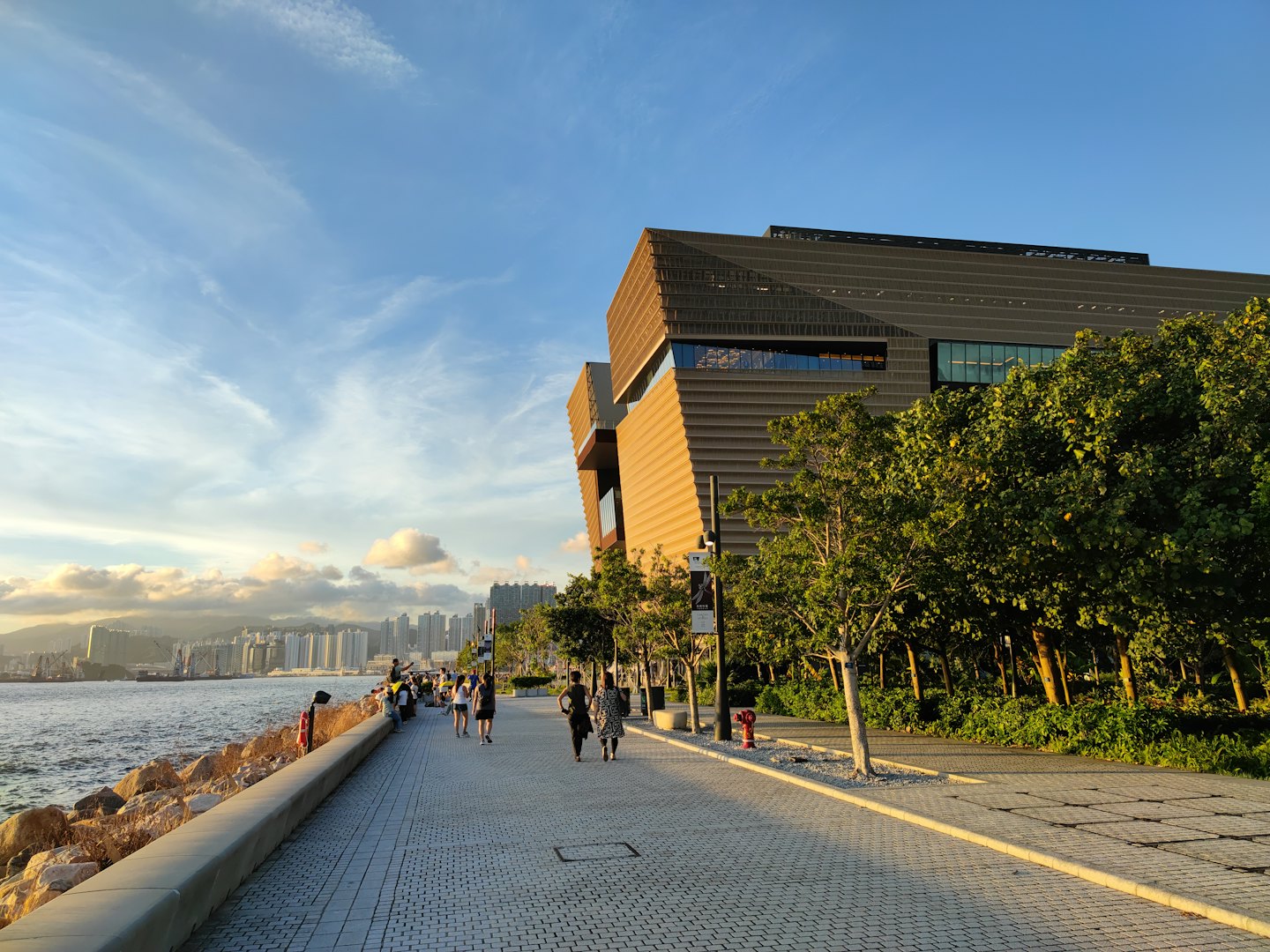 People walk by a modern building on a sunny waterfront promenade.