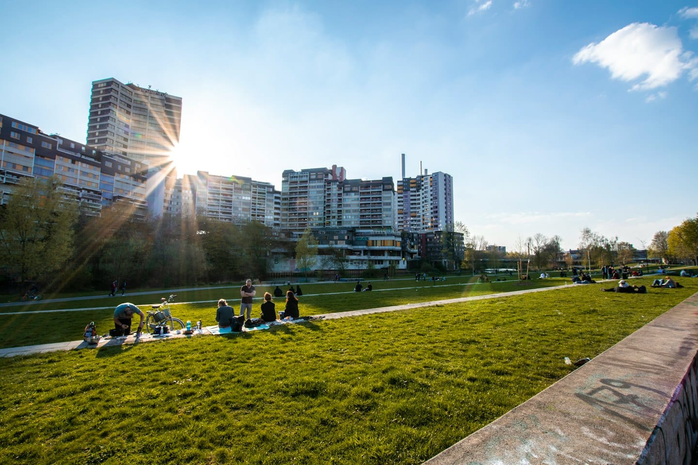 People relax on park grass near buildings under a sunny sky.